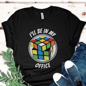 May include: Black t-shirt with a Rubik's Cube graphic inside a white circle, and the text "I'll be in my office". The shirt is paired with blue jeans and sneakers.
