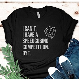 May include: Black t-shirt with the text "I CAN'T. I HAVE A SPEEDCUBING COMPETITION. BYE." and a white outline of a Rubik's Cube. The shirt is paired with blue jeans and white-soled sneakers.