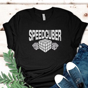 May include: Black t-shirt with the word "SPEEDCUBER" in white block letters above three white Rubik's Cube graphics. The shirt is paired with blue jeans and white-soled sneakers. A sprig of green leaves and a black circle with the word "black" are also in the image.