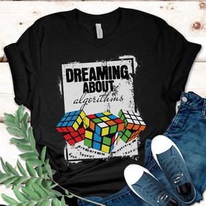 May include: Black t-shirt with the text "DREAMING ABOUT algorithms" and three Rubik's Cube graphics. The shirt is paired with blue jeans and navy blue sneakers with white soles. A sprig of green leaves is on the left.