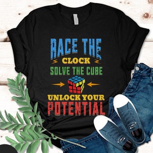 May include: Black t-shirt with a colorful graphic design featuring a Rubik's Cube and the text "Race the Clock Solve the Cube Unlock Your Potential".