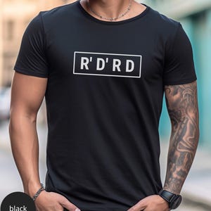 May include: Black crew neck t-shirt with the text "R' D' R D" in a white rectangular box. The shirt is a solid black color and has short sleeves. The person is wearing a watch and bracelet.
