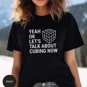 May include: Black t-shirt with the text "YEAH OK LET'S TALK ABOUT CUBING NOW" and a white graphic of a Rubik's Cube. The shirt is a crew neck style.