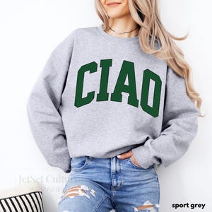 May include: A gray sweatshirt with the word "CIAO" printed in green letters. The sweatshirt is being modeled by a person wearing blue jeans.