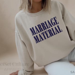 May include: A sand-colored sweatshirt with the text "MARRIAGE MATERIAL" in navy blue block letters. The sweatshirt is a crew neck style and appears to be made of a soft material. The words "JetSet Culture Apparel" are at the bottom left.