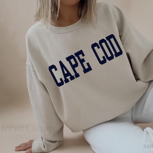 May include: Sand-colored sweatshirt with the text "CAPE COD" in navy blue block letters. The sweatshirt is a crew neck style and appears to be made of a soft material. The word "sand" is printed in the bottom right corner.