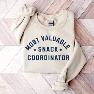 May include: A cream-colored sweatshirt with the text "MOST VALUABLE SNACK COORDINATOR" in navy blue, arched above and below a star. The sleeves are rolled up. The Gildan brand tag is visible.