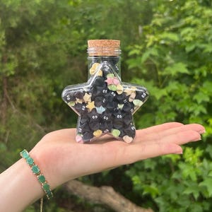 May include: A clear, star-shaped glass bottle filled with origami stars. The origami stars are black with cartoon eyes and colorful accents. The bottle has a cork stopper. A green bracelet is visible.