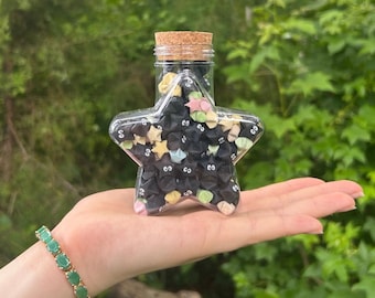 May include: A clear, star-shaped glass bottle filled with origami stars. The origami stars are black with cartoon eyes and colorful accents. The bottle has a cork stopper. A green bracelet is visible.