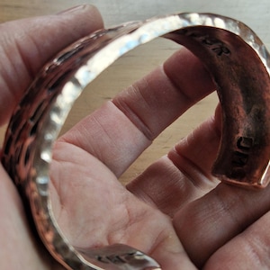 May include: A hammered copper cuff bracelet with a textured surface. The bracelet has a curved shape and is held in a hand. The cuff is a warm copper color and appears to be handmade. The letters "JM" are visible on the inside of the cuff.