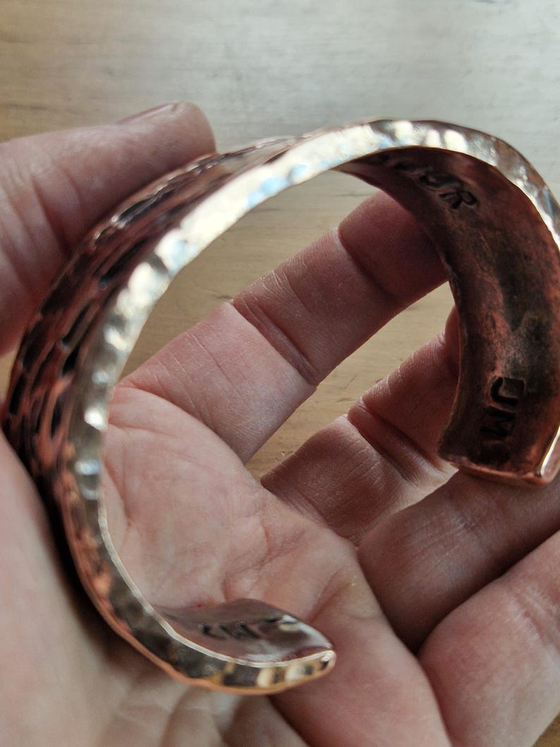 May include: A hammered copper cuff bracelet with a textured surface. The bracelet has a curved shape and is held in a hand. The cuff is a warm copper color and appears to be handmade. The letters "JM" are visible on the inside of the cuff.