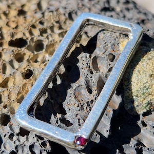 May include: A silver-toned rectangular pendant with a small, round, red gemstone set in one corner. The pendant is displayed on a textured, light brown and gray stone surface. The pendant's design is simple and modern.