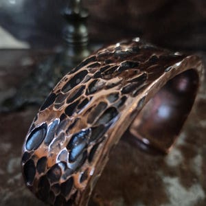 May include: A hammered copper cuff bracelet. The metal has a textured, irregular surface with dark and light brown tones. The open cuff design allows for easy wear. The bracelet is a statement piece.