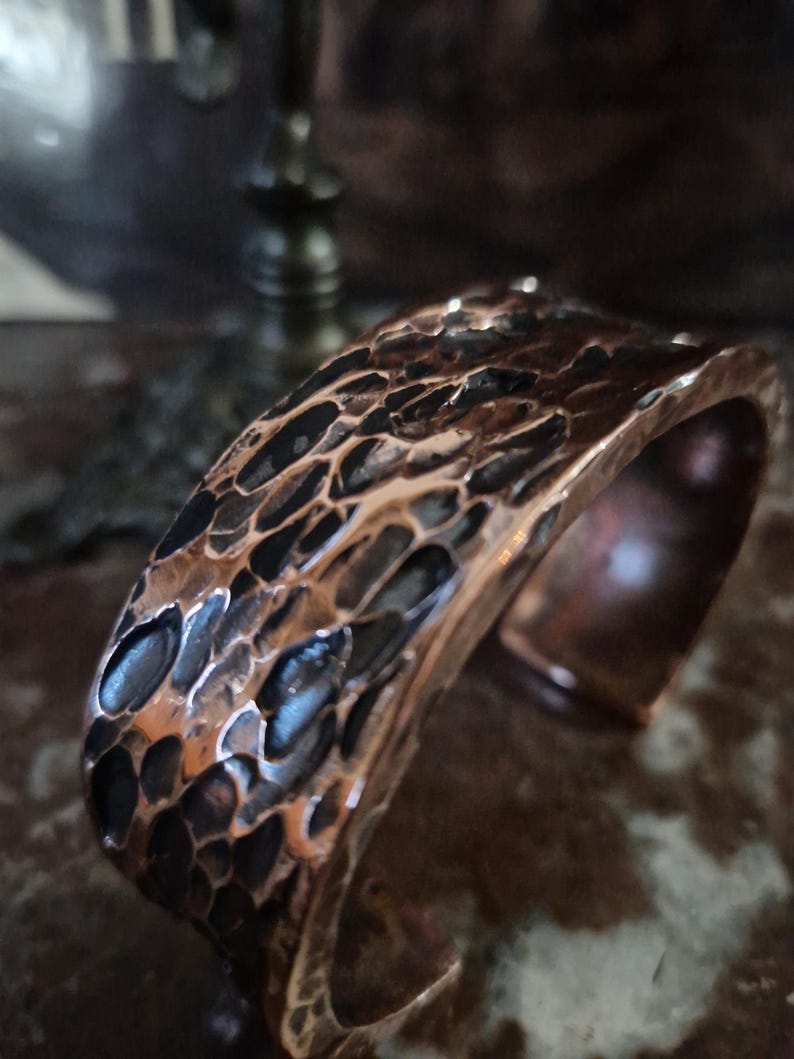 May include: A hammered copper cuff bracelet. The metal has a textured, irregular surface with dark and light brown tones. The open cuff design allows for easy wear. The bracelet is a statement piece.