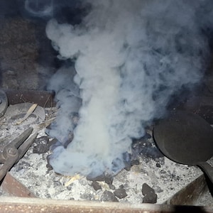 May include: A blacksmith's forge with tools and smoke. The image shows a forge with a bed of ash and charcoal, with tools like tongs and a round pan. A large plume of white smoke rises from the center, indicating recent activity.