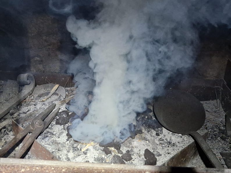 May include: A blacksmith's forge with tools and smoke. The image shows a forge with a bed of ash and charcoal, with tools like tongs and a round pan. A large plume of white smoke rises from the center, indicating recent activity.