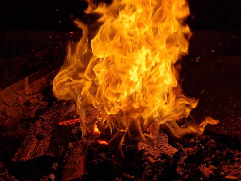 May include: A close-up shot of a vibrant fire with bright orange and yellow flames. The flames are rising from a bed of dark, charred wood and embers, creating a warm and intense visual.