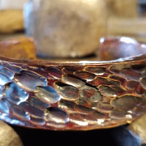 May include: A hammered copper cuff bracelet with a textured, scale-like pattern. The open-ended bracelet has a warm, reddish-brown color with hints of blue and silver. The cuff is approximately 1 inch wide.