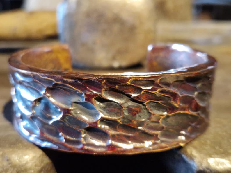 May include: A hammered copper cuff bracelet with a textured, scale-like pattern. The open-ended bracelet has a warm, reddish-brown color with hints of blue and silver. The cuff is approximately 1 inch wide.