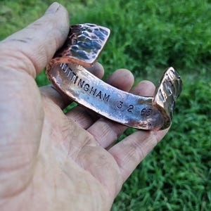 May include: Copper cuff bracelet with textured surface and the engraved text "NOTTINGHAM 32 65". The open-ended bracelet is held in a hand against a blurred green background.