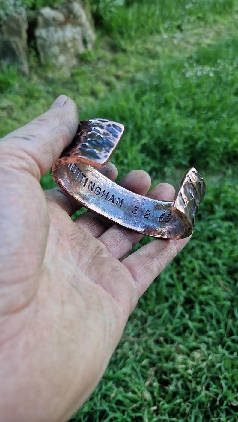 May include: Copper cuff bracelet with textured surface and the engraved text "NOTTINGHAM 32 65". The open-ended bracelet is held in a hand against a blurred green background.