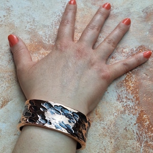 May include: A wide copper cuff bracelet with a hammered texture. The bracelet is worn on a wrist, showcasing its metallic sheen and unique design. The cuff is approximately 2 inches wide.