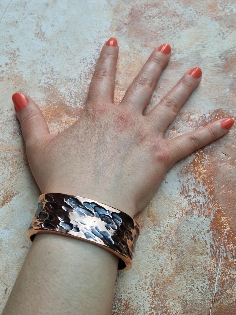 May include: A wide copper cuff bracelet with a hammered texture. The bracelet is worn on a wrist, showcasing its metallic sheen and unique design. The cuff is approximately 2 inches wide.