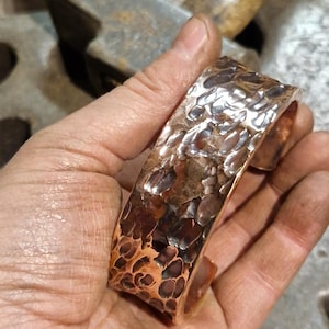 May include: A hammered copper cuff bracelet with a textured surface. The cuff is held in a hand, showcasing the intricate details and the warm tones of the metal. The cuff is approximately 2 inches wide.
