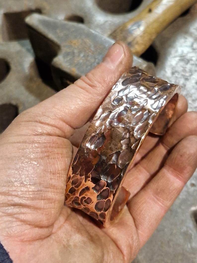 May include: A hammered copper cuff bracelet with a textured surface. The cuff is held in a hand, showcasing the intricate details and the warm tones of the metal. The cuff is approximately 2 inches wide.