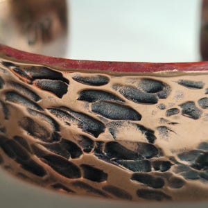 May include: Close-up of a copper cuff bracelet with a hammered texture. The bracelet has a reddish-brown interior and a textured surface with dark, irregular patterns. The cuff is open-ended and appears to be handmade.