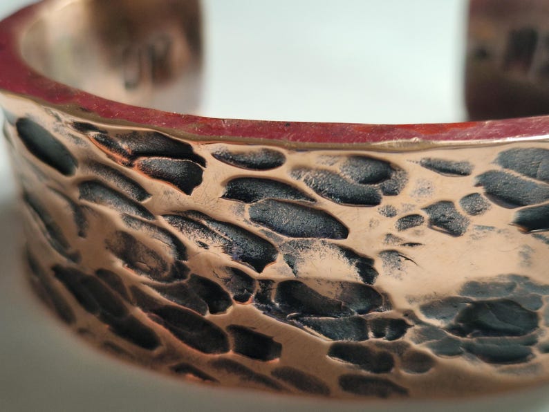 May include: Close-up of a copper cuff bracelet with a hammered texture. The bracelet has a reddish-brown interior and a textured surface with dark, irregular patterns. The cuff is open-ended and appears to be handmade.