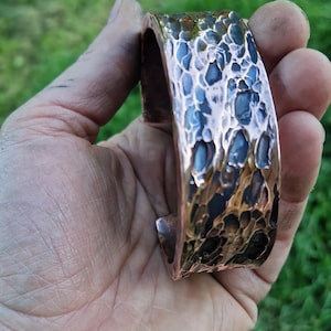 May include: Copper cuff bracelet with a textured, hammered surface. The bracelet has a wide band with a dark blue and bronze color scheme. The cuff is held in a hand against a blurred green background.