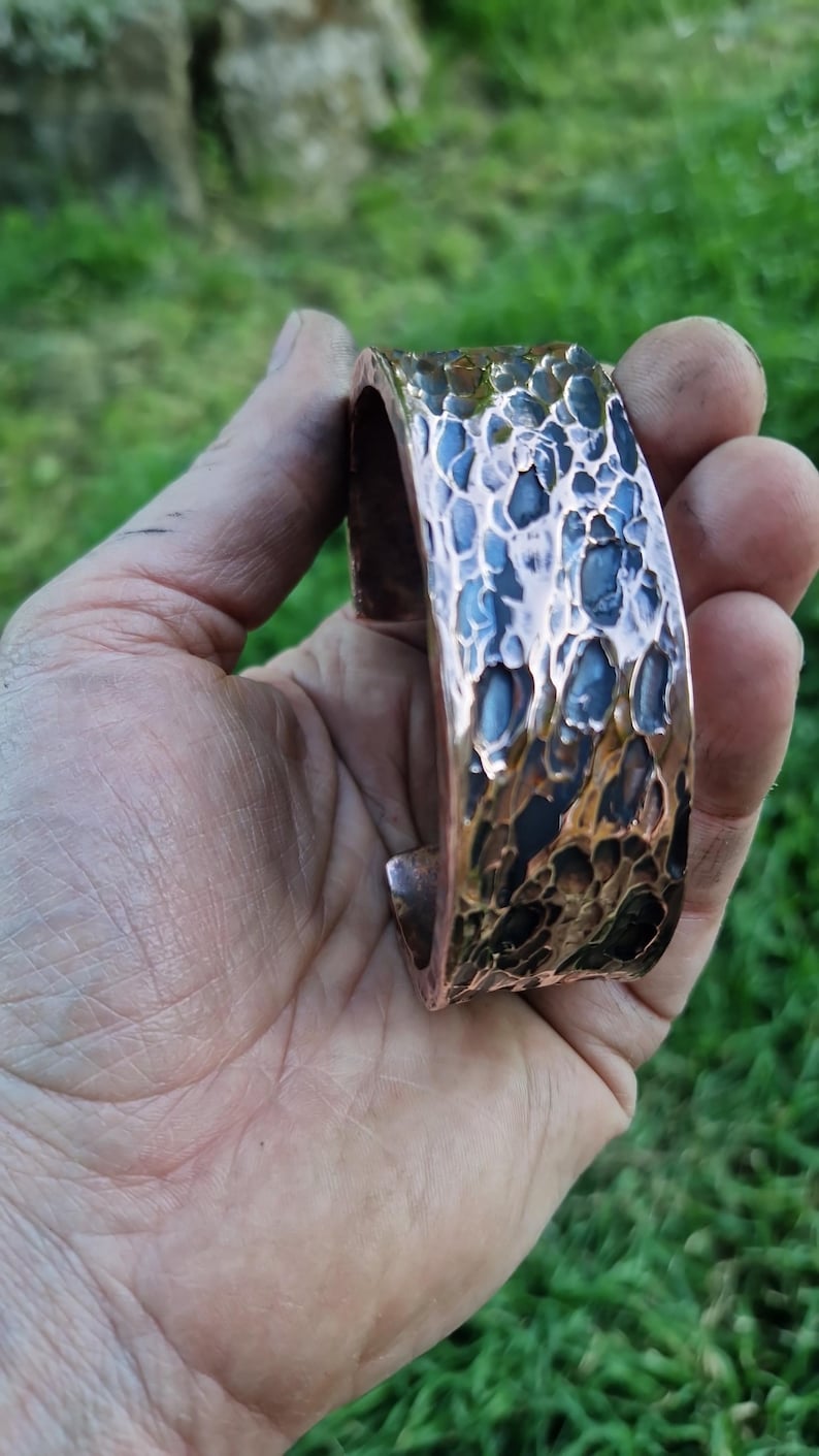 May include: Copper cuff bracelet with a textured, hammered surface. The bracelet has a wide band with a dark blue and bronze color scheme. The cuff is held in a hand against a blurred green background.