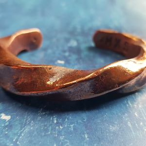 May include: A twisted copper bracelet with a hammered finish. The bracelet is open and has a slightly curved shape. The inside of the bracelet is marked with the word "MAX".