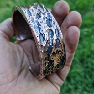 May include: A copper cuff bracelet with a textured, hammered surface. The bracelet has a dark, mottled finish with shades of blue, black, and brown. The open-ended design allows for easy wear. The bracelet is held in a hand against a blurred green background.