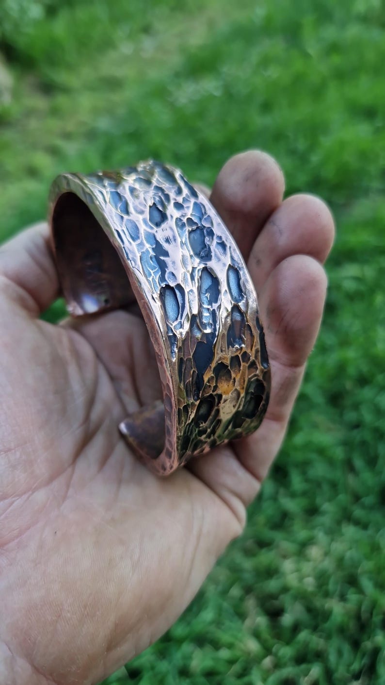 May include: A copper cuff bracelet with a textured, hammered surface. The bracelet has a dark, mottled finish with shades of blue, black, and brown. The open-ended design allows for easy wear. The bracelet is held in a hand against a blurred green background.