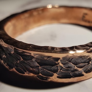 Pode incluir: Uma pulseira de cobre martelada com uma superfície texturizada e uma pátina preta. O interior da pulseira é gravado com as iniciais "E.J.M."