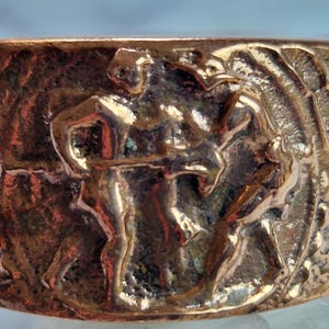 May include: A bronze-colored ring with a detailed relief sculpture. The sculpture depicts two figures in a dynamic pose, possibly engaged in a dance or ritual. The ring's surface has a textured, aged appearance, suggesting an antique or vintage style.