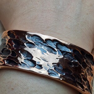 May include: A hammered copper cuff bracelet. The wide bracelet has a textured surface with dark and light brown tones. The cuff is worn on a wrist, showcasing the bracelet's design and craftsmanship. The metal reflects light, highlighting the hammered details.