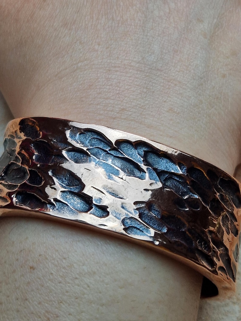 May include: A hammered copper cuff bracelet. The wide bracelet has a textured surface with dark and light brown tones. The cuff is worn on a wrist, showcasing the bracelet's design and craftsmanship. The metal reflects light, highlighting the hammered details.