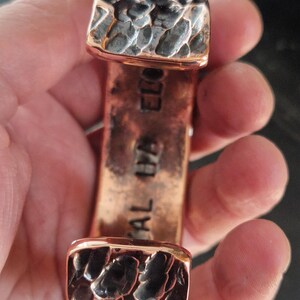 May include: A copper cuff bracelet with textured, dark gray accents on the ends. The bracelet has the words "CAL IYA ELO" stamped on the copper. The cuff is held in a hand.