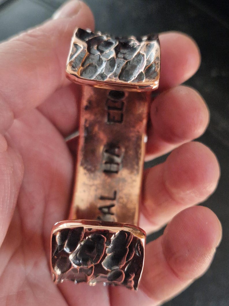 May include: A copper cuff bracelet with textured, dark gray accents on the ends. The bracelet has the words "CAL IYA ELO" stamped on the copper. The cuff is held in a hand.