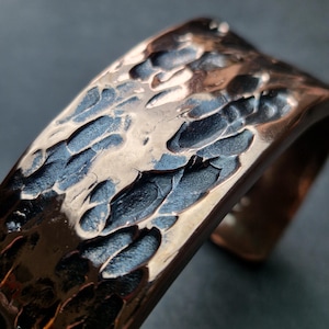 May include: Close-up of a hammered copper cuff bracelet. The metal has a textured surface with dark blue and bronze tones. The cuff is open on one side.