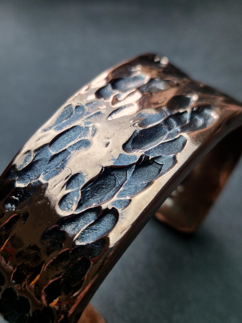 May include: Close-up of a hammered copper cuff bracelet. The metal has a textured surface with dark blue and bronze tones. The cuff is open on one side.