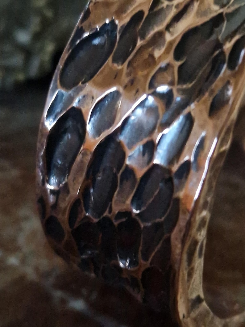 May include: Close-up of a copper-colored metal ring with a textured surface. The ring's surface has a pattern of irregular, dark-colored indentations, creating a unique, organic appearance. The metal reflects light, highlighting the texture.