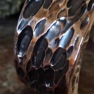 May include: Close-up of a copper-colored metal ring with a textured surface. The ring's surface has a pattern of irregular, dark-colored indentations, creating a unique, organic appearance. The metal reflects light, highlighting the details of the design.