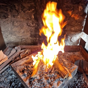 May include: A close-up shot of a blacksmith's forge with a bright orange and yellow fire burning. The fire is fueled by wooden logs in a metal container, surrounded by ash and embers. The background is a stone wall.