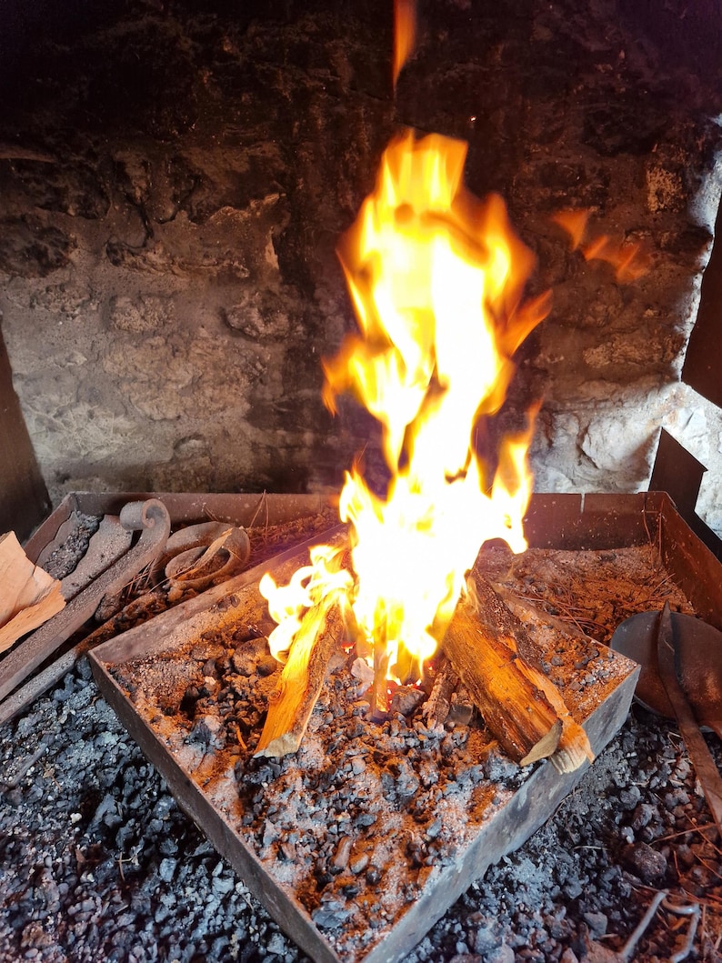 May include: A close-up shot of a blacksmith's forge with a bright orange and yellow fire burning. The fire is fueled by wooden logs in a metal container, surrounded by ash and embers. The background is a stone wall.