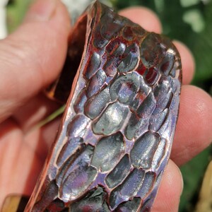 May include: Close-up of a copper cuff bracelet with a textured, scale-like pattern. The bracelet has a dark, reddish-brown color with hints of purple and blue. The cuff is held in a person's hand.