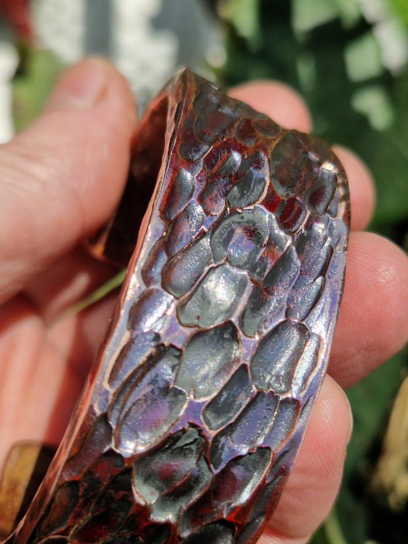 May include: Close-up of a copper cuff bracelet with a textured, scale-like pattern. The bracelet has a dark, reddish-brown color with hints of purple and blue. The cuff is held in a person's hand.